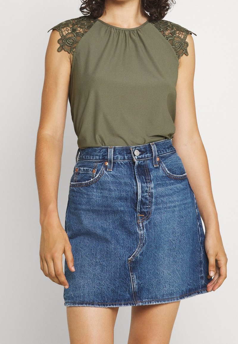 Femme portant un top sans manches vert olive avec des épaules en dentelle et une jupe en denim bleue taille haute avec des poches avant et une fermeture boutonnée.