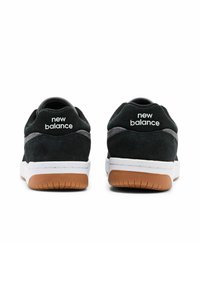 Zapatillas de ante negras con una media suela blanca y suela de goma color caramelo. Presentan un tacón redondeado y la marca "new balance" en la parte trasera.