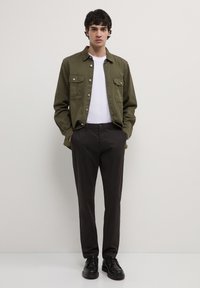 Camicia verde oliva a bottoni con due tasche sul petto, indossata sopra una maglietta bianca, abbinata a pantaloni neri e scarpe nere, sfondo neutro.