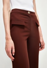 Detailní záběr na osobu v hnědých kalhotách s vysokým pasem, které mají kapsy s klopami a zlatý knoflík, doplněné bílým topem bez rukávů.