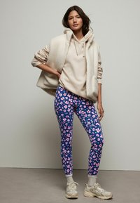 Beige fleece vest over een lichtbeige hoodie, gecombineerd met blauwe leggings met roze bloemenpatronen en grijze sneakers met witte accenten.