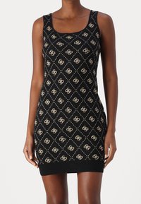Femme portant une robe en maille noire sans manches avec un motif de losanges beiges et de petits logos répartis sur toute la surface.