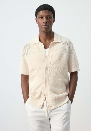 Jeune homme portant une chemise en maille crème à manches courtes boutonnée sur un haut blanc, les mains dans les poches d’un pantalon blanc.