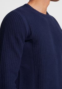 Pull en maille côtelée bleu marine avec col rond et motif texturé, présentant des designs en maille contrastés le long des côtés.