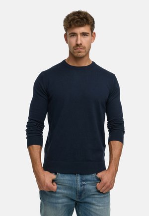 Mand med kort krøllet hår iført en navyblå sweater og lyseblå jeans, stående med hænderne i lommerne foran en ensfarvet baggrund.