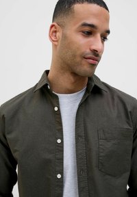 Camicia button-up verde scuro con taschino sul petto, bottoni bianchi e una texture sottile, indossata sopra una maglietta a girocollo grigia chiara.