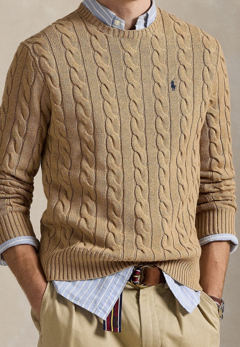 Homme portant un pull en maille torsadée beige par-dessus une chemise bleue rayée rentrée dans un pantalon beige, mains dans les poches.