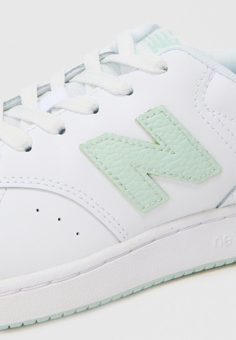 Zapatilla blanca con parte superior de cuero liso, que presenta un logotipo "N" texturizado en verde menta. Diseño de cordones y acentos en azul claro en la lengüeta.