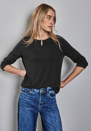 Maglietta nera a maniche lunghe con scollatura rotonda e dettaglio a occhiello, abbinata a jeans in denim blue con un taglio classico e accenti di cucitura.