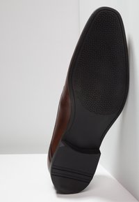 Bruine leren nette schoen met zwarte rubberen zool, zichtbaar met gestructureerde profiel en gestapelde hak tegen een witte achtergrond.