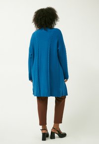 Cardigan lungo blu con lavorazione a maglia testurizzata, caratterizzato da motivi a treccia e bordi a costine, abbinato a pantaloni slim-fit marroni e scarpe nere con tacco a blocco.