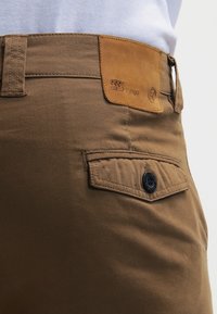 Pantaloni di cotone marroni con finitura testurizzata, caratterizzati da una toppa in pelle con logo in rilievo e un dettaglio di tasca laterale con bottone.