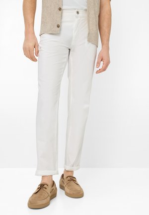 Mand iført hvide optrukne bukser, brune snørestøvler til sejlads og en beige ærmeløs cardigan over en hvid skjorte.