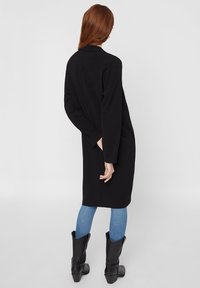 Černý dlouhý kabát s dlouhými rukávy, volný střih, středně dlouhý lem, nošený přes světle modré skinny džíny a černé kozačky po kolena.