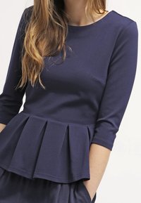 Granatowa bluzka peplum z rękawami o długości trzech ćwierć. Gładki materiał, plisowany krój w talii, okrągły dekolt oraz detal w postaci pojedynczej kieszonki na piersi.