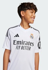 Hvit fotballtrøye med svarte detaljer og korte ermer, med "Emirates FLY BETTER" trykt på forsiden og en logo på brystet.