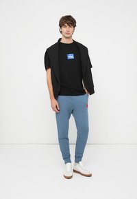 Černé tričko s modrým logem, vrstvené pod černou bundou; šedo-modré tepláky s červeným akcentem; bílé tenisky s hnědou podrážkou.