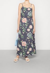 Blommig maxiklänning med spagettiband, mörk marinblått tyg, stor rosa och grön blommönster, kombinerad med vita slip-on skor och en ljusblå väska.