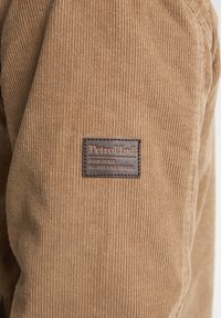 Tessuto di velluto a coste marrone con righe verticali; presenta una toppa rettangolare in pelle con "Petrol Ind" e "DENIM DRIVEN" impressi in oro.