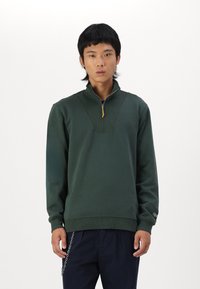 Sudadera verde de media cremallera hecha de una tela suave, con acentos acanalados y una etiqueta amarilla. Mangas largas y corte holgado.