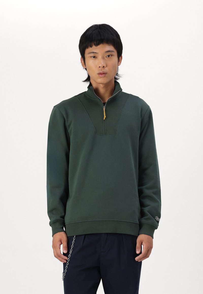Sudadera verde de media cremallera hecha de una tela suave, con acentos acanalados y una etiqueta amarilla. Mangas largas y corte holgado.