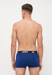 Pantalones cortos tipo boxers azul marino con una cinturilla negra que presenta el logo de Lacoste. Tela suave con una forma ajustada y sin patrones visibles.