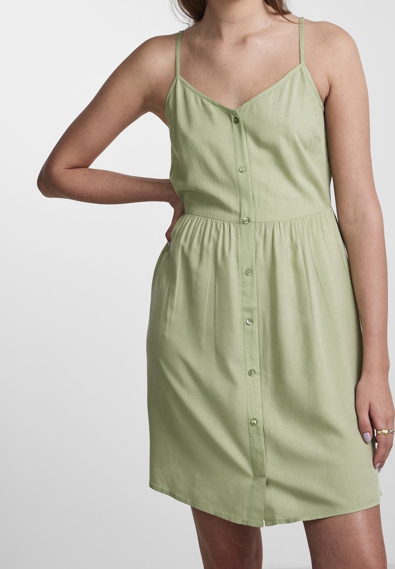 Femme portant une robe sans manches vert clair à boutons sur le devant avec de fines bretelles, une main sur la hanche, sur fond blanc uni.