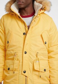 Geel gewatteerde parka met een met bont gevoerde capuchon, twee voorzakken, een trekkoord in de taille en zwarte knopen. Heeft een gestructureerde voering.