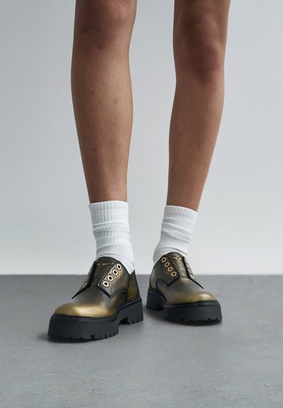 Metallisch goldene Schuhe mit rundem Zehenbereich und schwarzer Gummisohle, mit Ösen und einer strukturierten Oberfläche. Dazu werden weiße Knöchelsocken getragen.