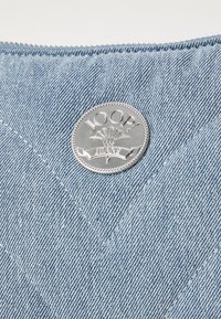 Denim tkanina v svetlo modri barvi z strukturirano površino; vsebuje okroglo srebrno kovinsko logotip z napisom "JOOP! JEANS" in dekorativnimi poudarki.