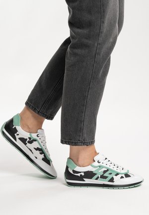 Personne portant des baskets à motifs noir et blanc avec des accents vert menthe et un jean noir coupé, debout sur un fond blanc.