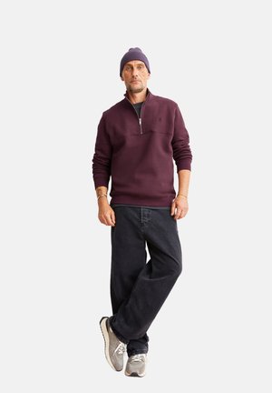 Burgunderfarbener Reißverschluss-Pullover mit einem schwarzen Unterhemd, kombiniert mit lockeren schwarzen Hosen und grauen Sneakers; das Modell trägt eine lila Mütze.