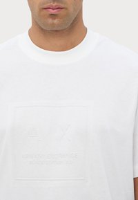 T-shirt in cotone bianco con un design del logo in rilievo che presenta "A|X" e "ARMANI EXCHANGE BLACK EDITION 2.0" al centro. Stile a girocollo.