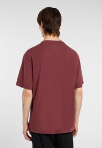 Osoba s krátkými hnědými vlasy, oblečená v volné vínové polokošili s krátkým rukávem a černých kalhotách, stojící zády k objektivu na jednobarevném pozadí.