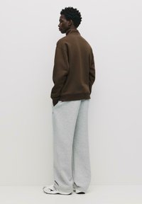 Hnedý sweatshirt s vysokým golierom v kombinácii so voľnými šedými teplákmi. Tkanina vyzerá byť jemná a outfit má uvoľnený strih a bočné vrecká.