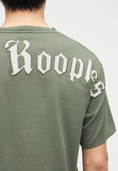 Osoba oblečená v olivovo zelenom tričku s veľkými bielymi gotickými písmenami "Kooples" cez hornú časť chrbta a plece.