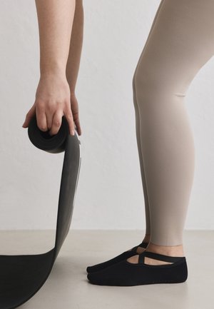 Person i beige leggings og sorte grebsokker ruller en sort yogamåtte ud på et lyst gulv mod en enkel væg.