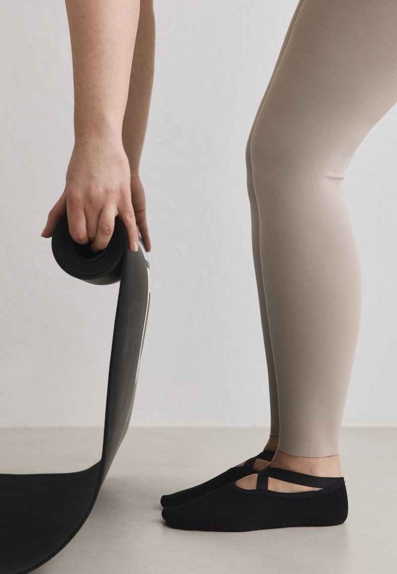 Personne en leggings beige et chaussettes antidérapantes noires déroulant un tapis de yoga noir sur un sol clair contre un mur uni.
