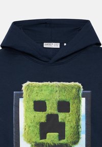 Námornícke modré mikina s grafickým motívom na tvári pokrytej trávou. Obsahuje okrúhlu kapucňu a mäkký bavlnený materiál. Štítok viditeľný vo vnútri.