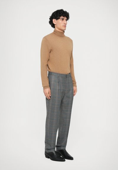 Brauner Rollkragenpullover kombiniert mit grauen karrierten Hosen. Die Hosen haben orangefarbene und blaue Akzente, eine taillierte Passform und schwarze Stiefel.