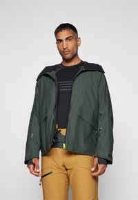 Chubasquero verde impermeable con capucha, con múltiples bolsillos y cremalleras, combinado con una camiseta térmica negra y pantalones beige.