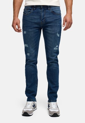 Mężczyzna ubrany w obcisłe, niebieskie, przetarte dżinsy oraz białe trampki, stojący na tle jednolitego jasnego tła.