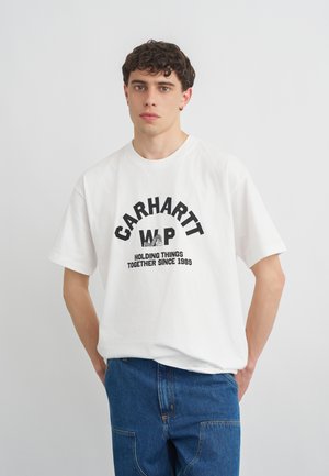 Młody mężczyzna z krótkimi kręconymi włosami, ubrany w białą koszulkę Carhartt z czarnym napisem i niebieskie dżinsy, stojący na jednolitym jasnym tle.