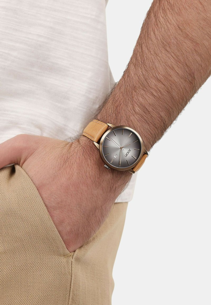 Runder silberfarbener Uhr mit grauem Zifferblatt, braunem Lederarmband und minimalistischem Stundenmarkierungen. Trägt die Uhr an einem Handgelenk mit einem hellen Stoff.