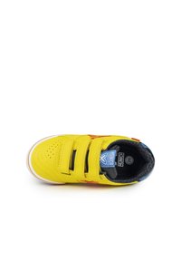 Zapatilla amarilla con tres correas de Velcro, parte superior de cuero texturizado, interior acolchado negro, acentos naranjas y azules, y suela de goma blanca.