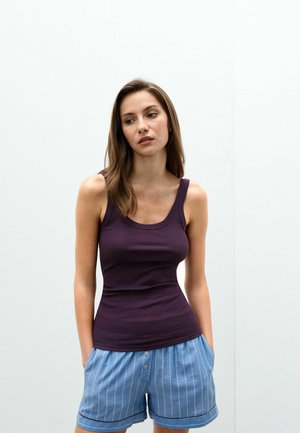Jonge vrouw met bruin haar kijkt opzij, draagt een mouwloos paars topje en blauw gestreepte korte broek met handen in de zakken tegen een witte achtergrond.