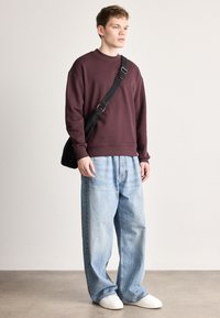 Bordó pulóver, bő szabású világoskék farmernadrág és fehér tornacipő. Fekete keresztpántos táskát tart, semleges háttér előtt áll.