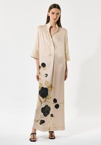 Femme portant une longue robe beige avec des motifs floraux noirs et bruns, des manches trois-quarts et des sandales noires à lanières sur un fond blanc.
