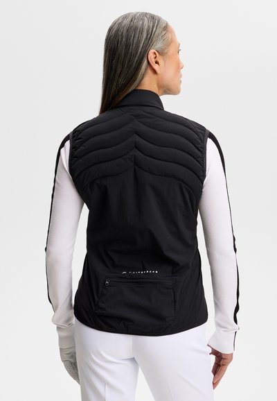 Kvinde med langt gråt hår iført en sort quiltet vest over en hvid langærmet top og hvide bukser, set bagfra mod en ensfarvet baggrund.