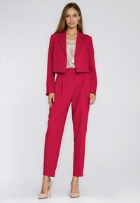 Różowy garnitur damski skrojony na miarę z krótką marynarką i spodniami z wysokim stanem. Pod spodem teksturowany top z cekinami. Całość dopełniają neutralne szpilki.